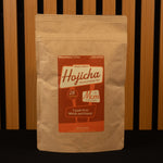 Kraft paper coffee bag with a red label featuring 'Hojicha' branding against a wooden slat background.